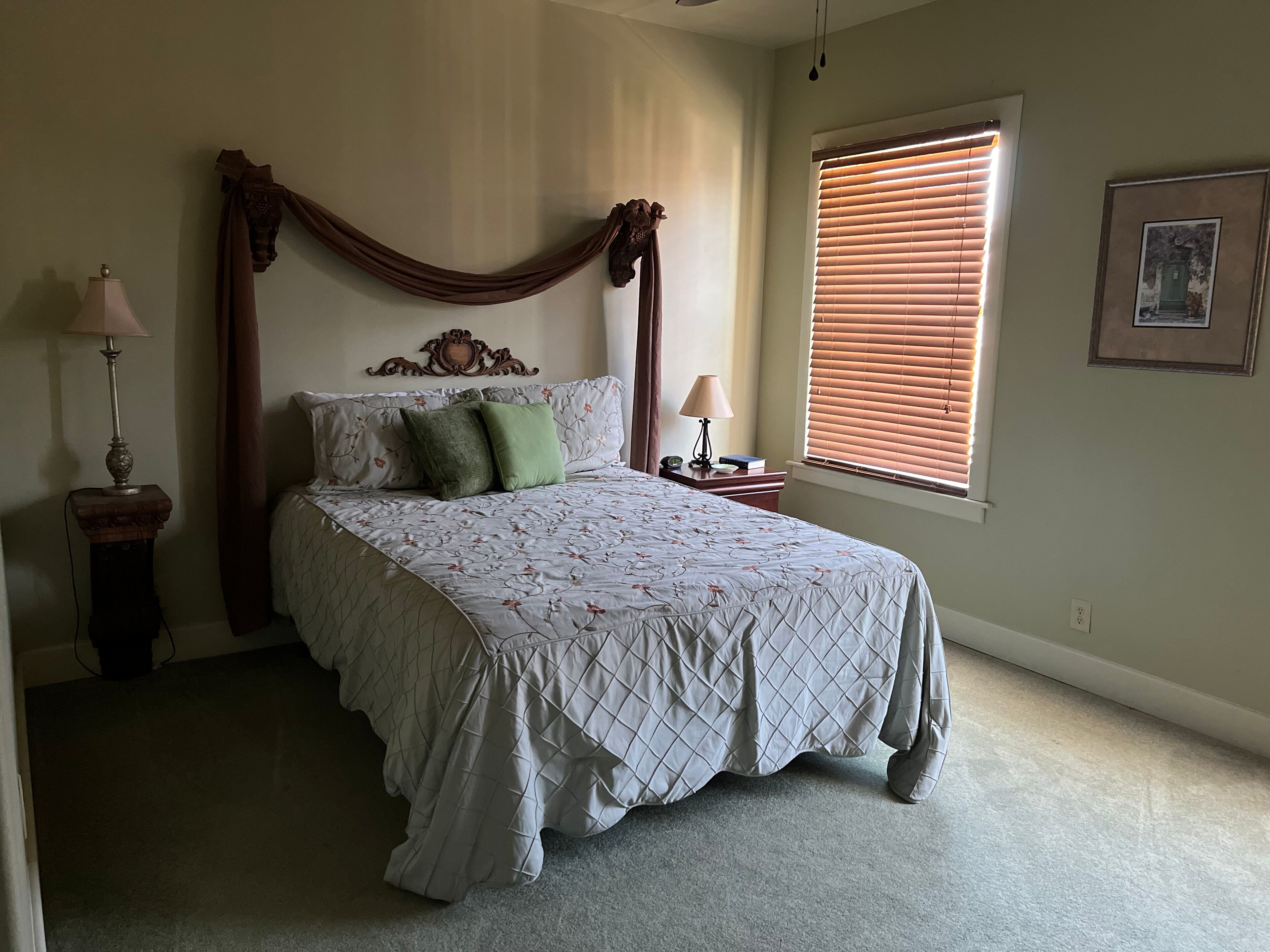 A bedroom with a bed, a nightstand, a lamp, a window with blinds, and a framed picture on the wall.