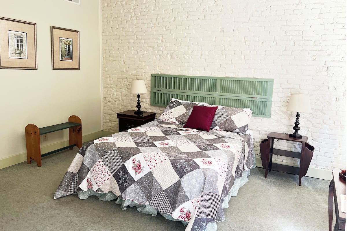A bedroom with a quilted bedspread, a green headboard, two bedside tables with lamps, and a bench against the wall.