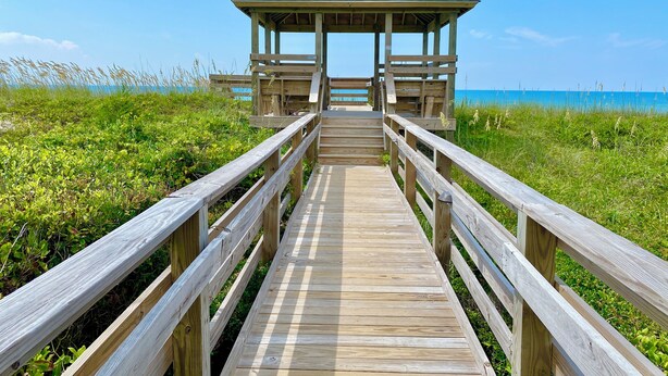 A wooden boardwalk leading to a gazebo with a sloped roof, surrounded by green vegetation and a clear blue sky. - Charming studio with Ocean View, AC, wifi, pool, on-site laundry room (Atlantic Beach)