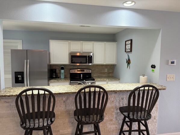 A modern kitchen with white cabinets, stainless steel appliances, and a granite countertop. There are three black bar stools in front of the counter.