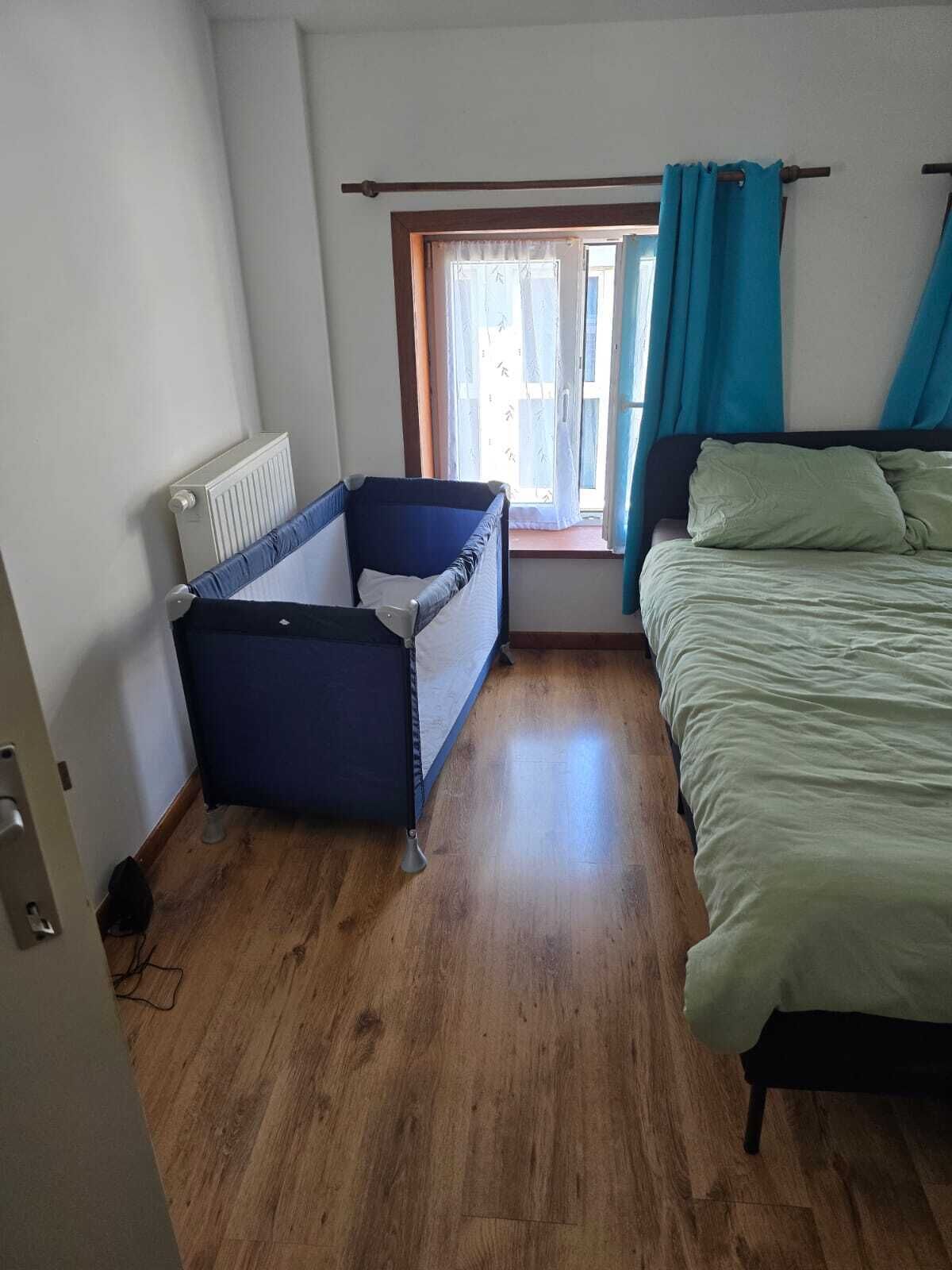 Een kamer met een houten vloer, een bed met groene beddengoed, een blauwe stoel en een raam met blauwe gordijnen.