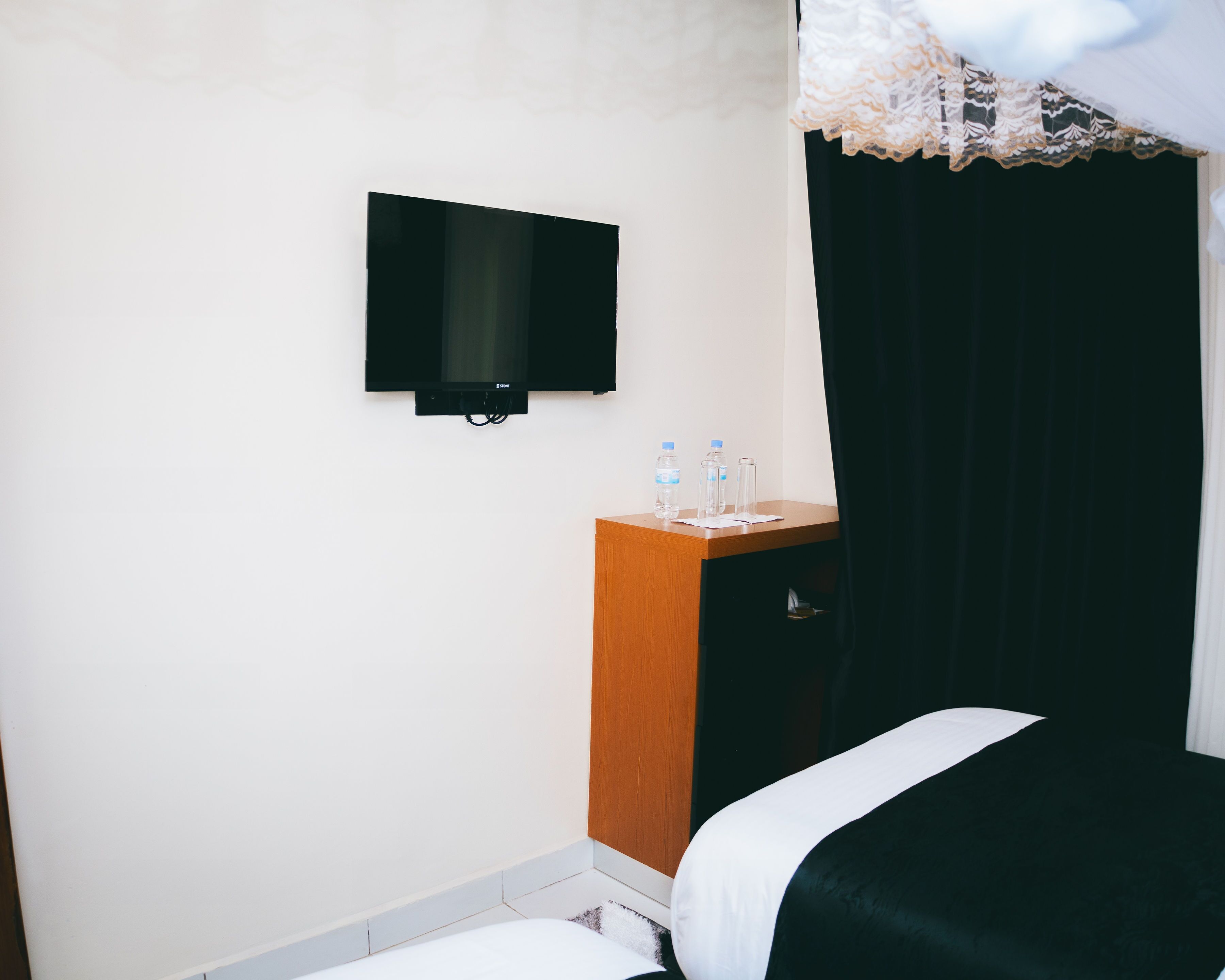 Una habitación de hotel con una cama, una mesita de noche de madera, un televisor montado en la pared y una ventana con cortina negra.
