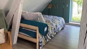 A loft bedroom with a sloped ceiling, a bed with patterned bedding, a wicker chair, and a view of greenery outside. - The little house in the village (Noyers)
