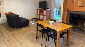 A living room with a wooden dining table, black chairs, a black sofa, a TV on a pink cabinet, and a fireplace. - The little house in the village (Noyers)