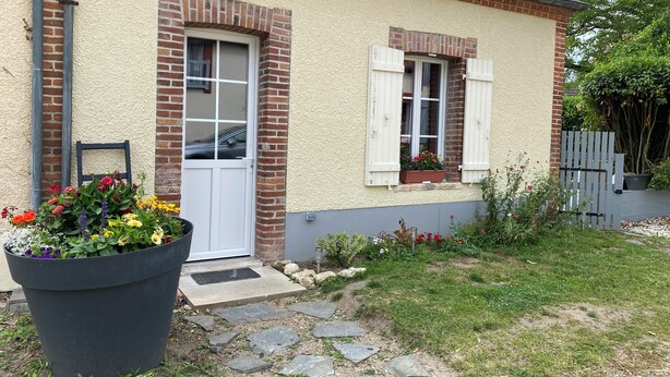 A house with a brick facade, a white door, and a window with shutters. There is a large planter with flowers in front of the door. - The little house in the village (Noyers)