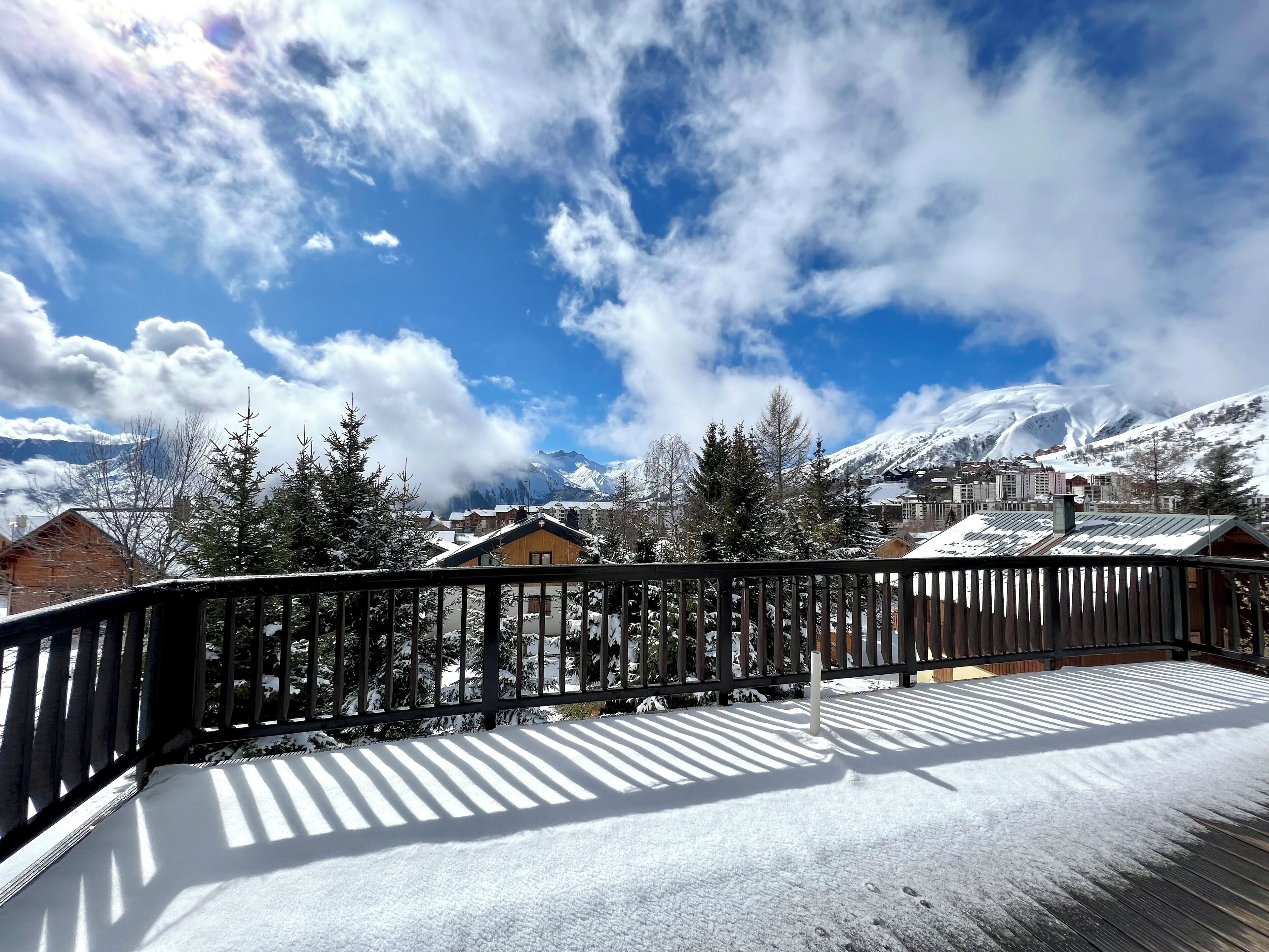 A snow-covered deck with a wooden railing, overlooking a town with buildings and snow-covered mountains in the background.
