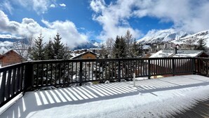 Un deck cubierto de nieve con baranda de madera, con vista a un pueblo y montañas nevadas al fondo.