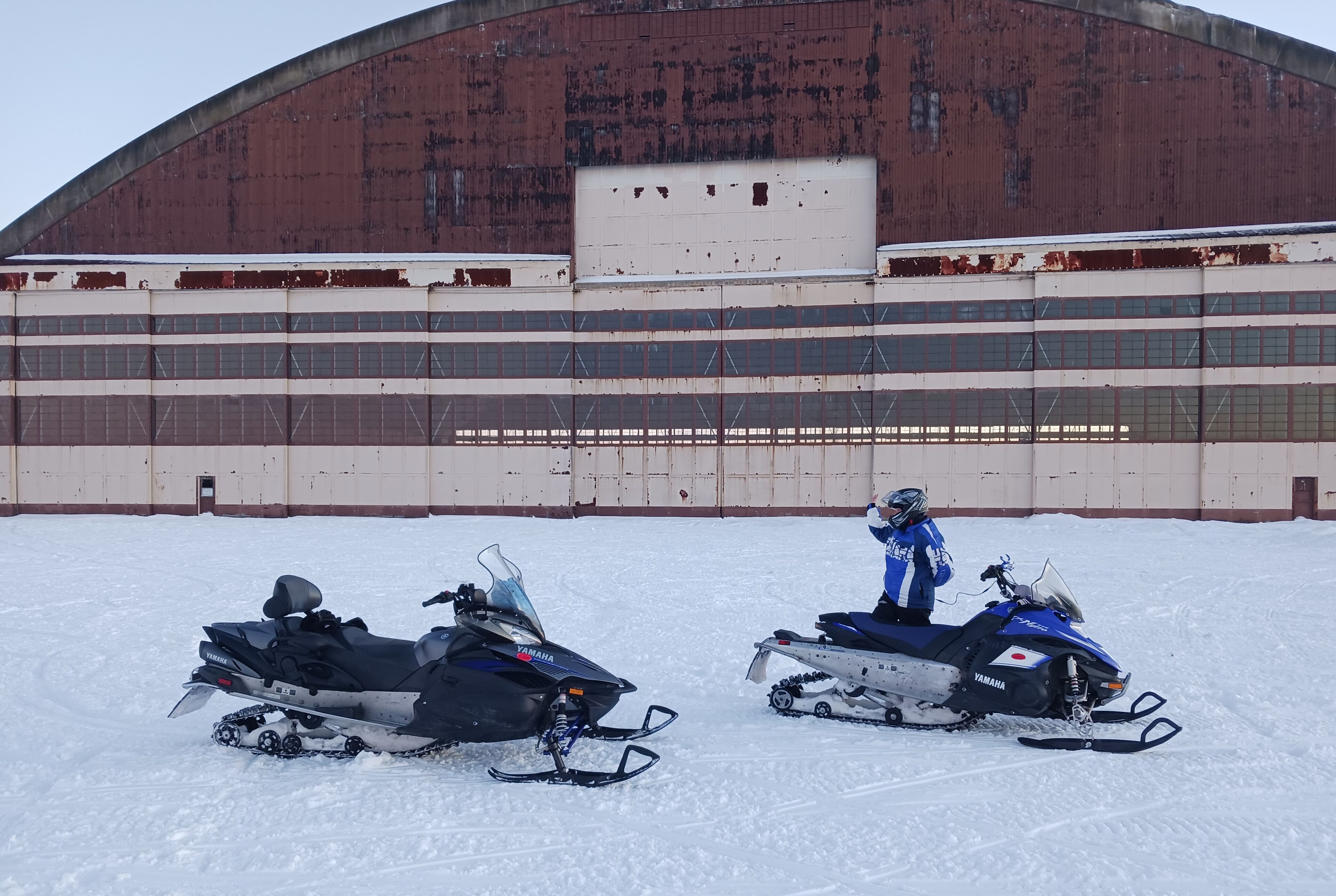 Dos motos de nieve estacionadas frente a un edificio grande de estilo industrial con techo curvo y varias ventanas.