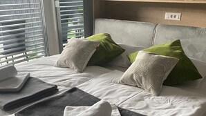 Een net opgemaakt bed met een wit-grijze deken, groene en witte kussens, en een opgevouwen witte handdoek. Daarboven bevindt zich een plank met decoratieve items en een kleine plant.