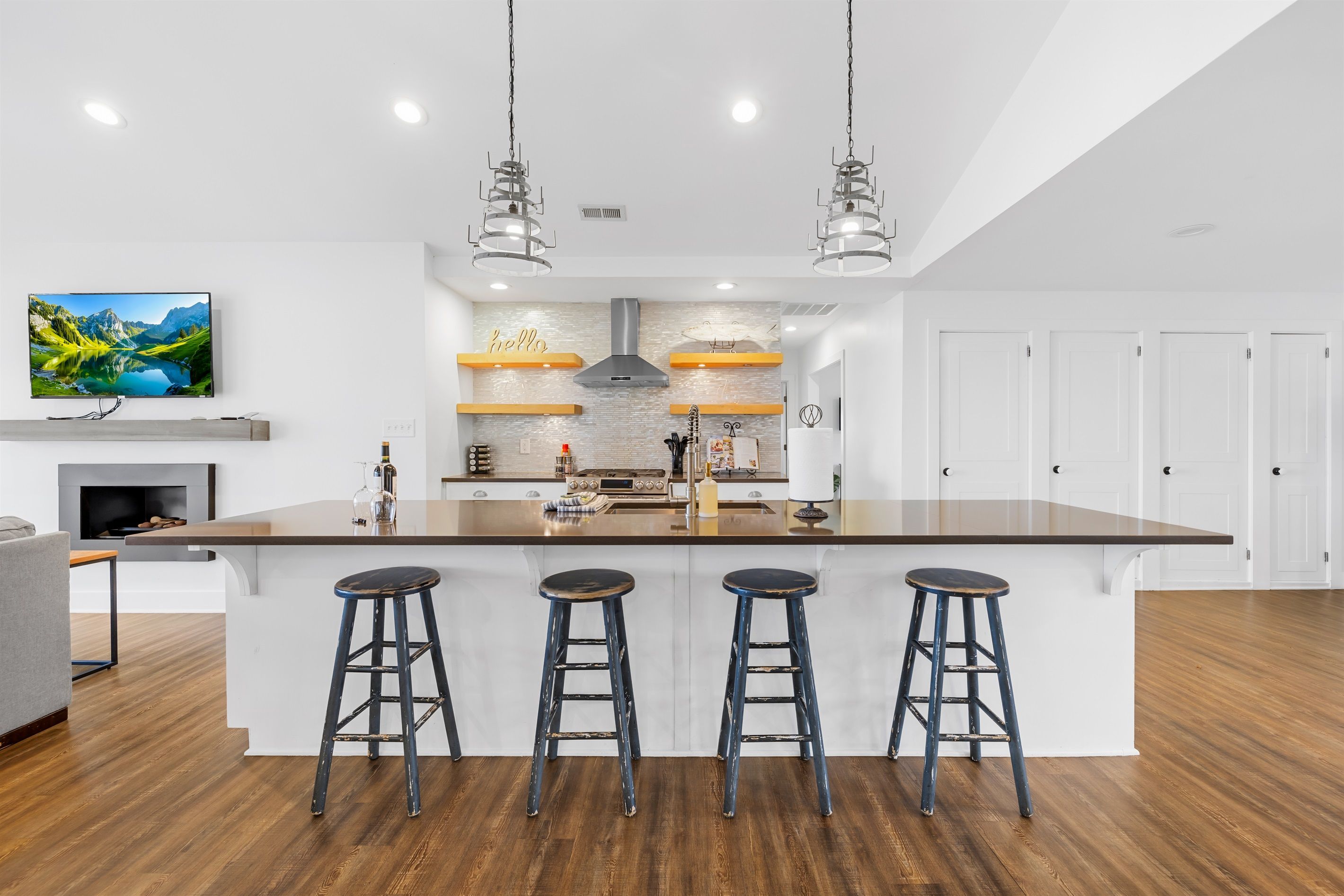 A modern kitchen with a central island, bar stools, pendant lights, and a TV on the wall.