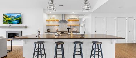 A modern kitchen with a central island, bar stools, pendant lights, and a TV on the wall.
