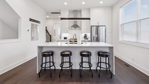 A modern kitchen with a central island, bar stools, stainless steel appliances, and a large window with blinds.