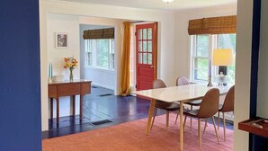 A room with a dining table, chairs, a red door, windows with curtains, a lamp, and a vase with flowers. - Tranquil farmhouse with WiFi in charming Cape Rosier (Brooksville)