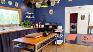 A kitchen with a wooden table, a stove, and hanging pots. There is a window with a view of trees, a washing machine, and a dog on a rug. - Tranquil farmhouse with WiFi in charming Cape Rosier (Brooksville)