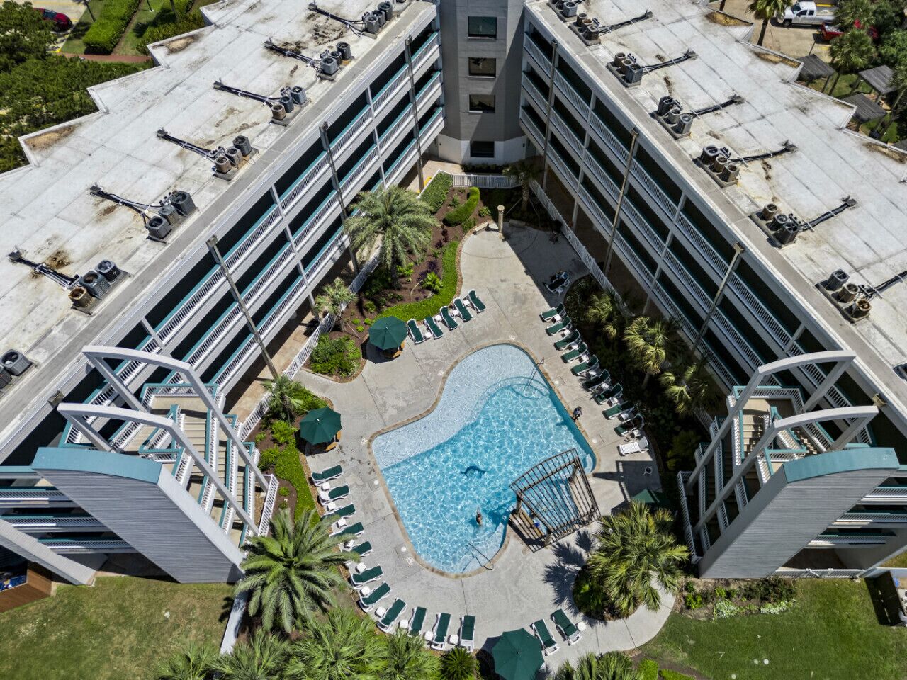 An aerial view of a modern hotel with a central swimming pool surrounded by lounge chairs and palm trees.