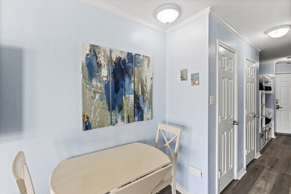 Una habitación con pared azul claro, una mesa color beige, una silla blanca y un cuadro abstracto de gran tamaño. Se observan puertas blancas y un estante con objetos decorativos.