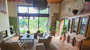 Ein großzügiger Wohnraum mit Kamin, bequemen Sitzgelegenheiten und großen Fenstern mit Blick auf den Garten.