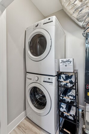 Een gestapelde wasmachine en droger met een bordje 'Wasserij', een rek met opgevouwen kleding en een metalen kast.
