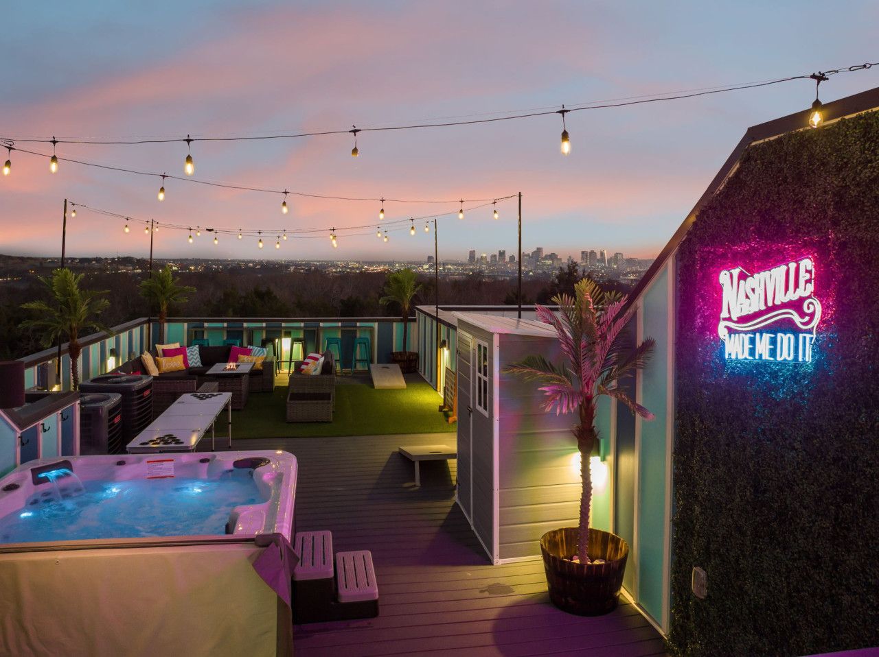 Een terras op het dak met een jacuzzi, buitenmeubels, lichtslingers en een neonbord.