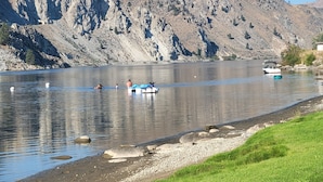 Un lago sereno con barcos, una orilla cubierta de hierba y montañas rocosas al fondo.