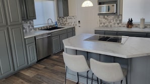 Una cocina moderna con armarios grises, una isla grande con taburetes de bar, un microondas y una ventana con persianas.
