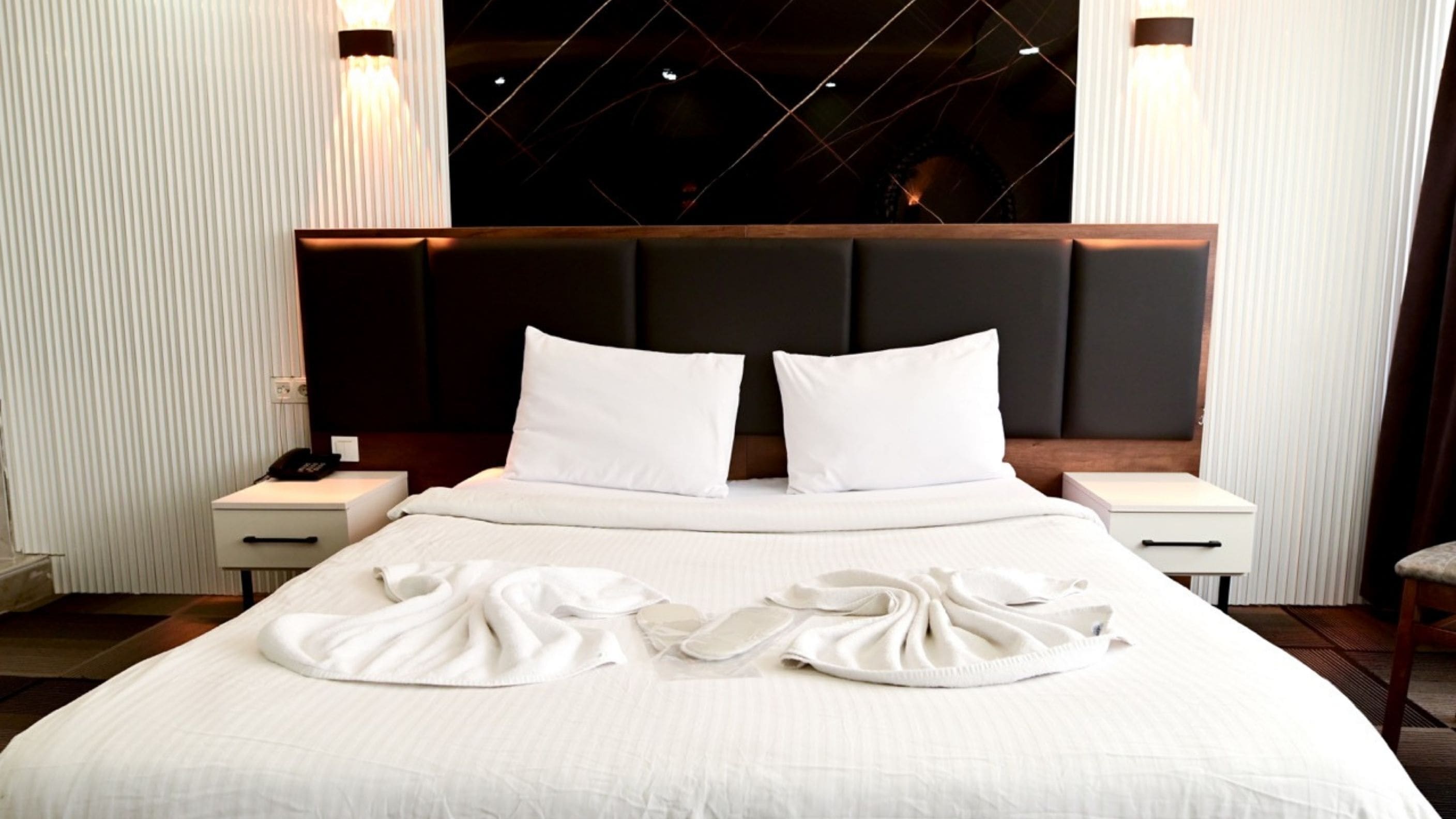 A neatly made bed with white linens and pillows, two bedside tables with a phone and a lamp, and a dark headboard.