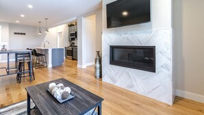 Una cocina moderna con microondas integrado, un comedor con mesa de madera y taburetes de bar, y una chimenea con televisor montado arriba.
