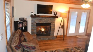 A living room with a fireplace, a television mounted above it, a sofa with patterned cushions, and a bookshelf. - Locust Flats - PET-FRIENDLY MOUNTAIN Cabin = Decks-Views-Firepit (Franklin)