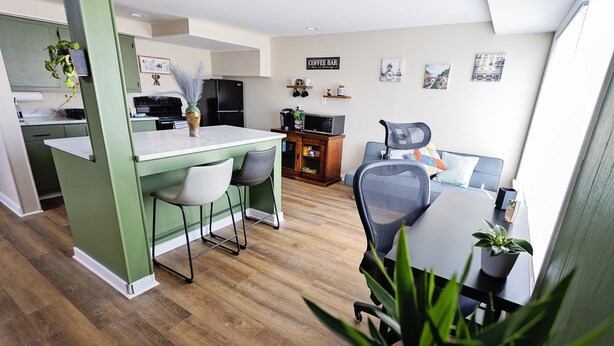 Una cocina moderna con armarios verdes, un comedor con mesa y sillas, y una sala de estar con sofá y cartel de bar de café.