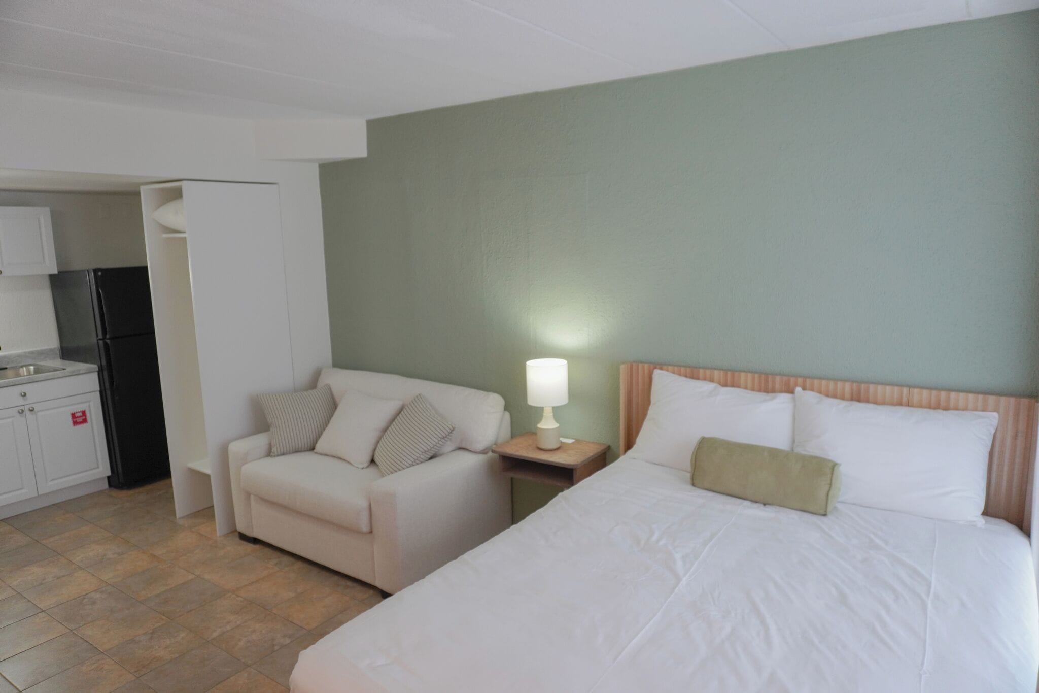 Hotelkamer met een bed, een stoel, een nachtkastje met een lamp, een kitchenette en een groene wand.