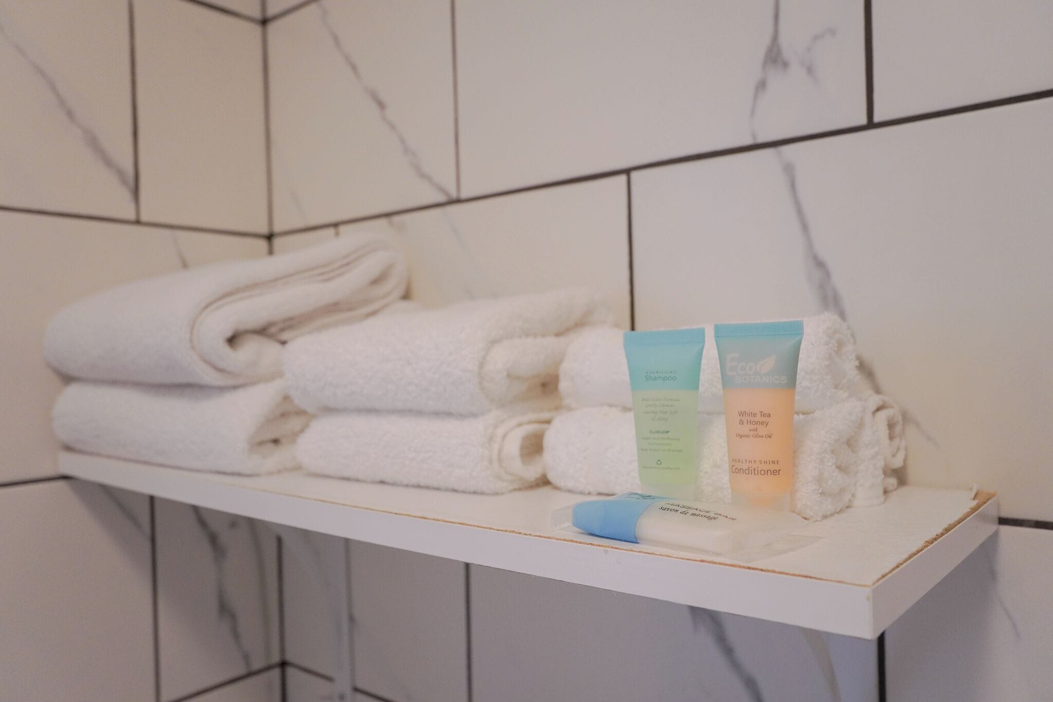 A bathroom with a white marble wall and a shelf holding folded towels and two bottles of shampoo and conditioner.
