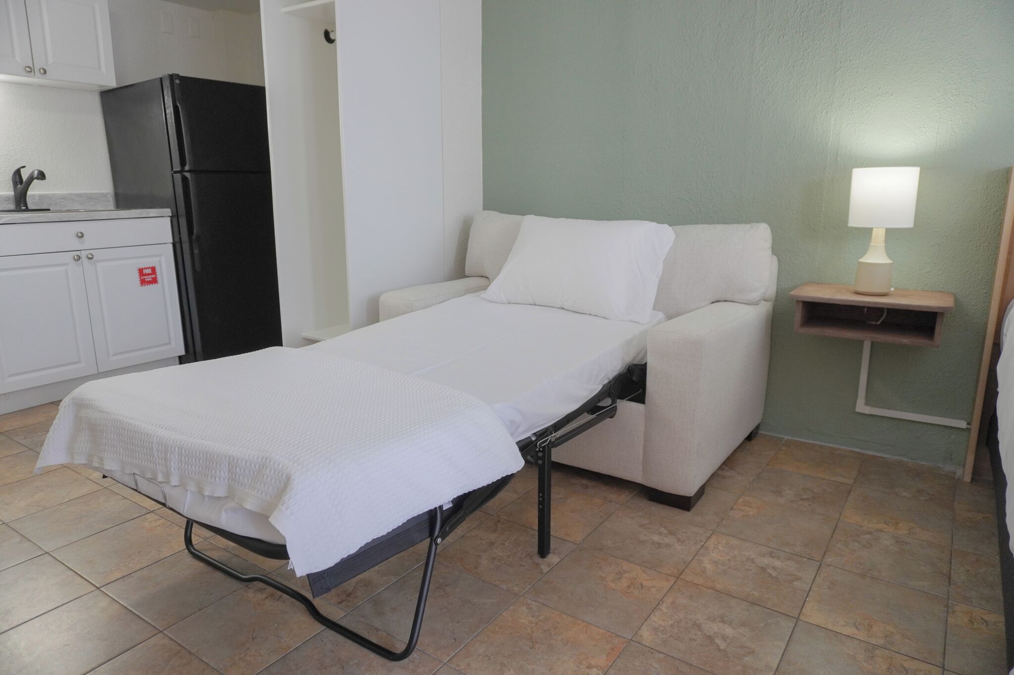 A single bed with a white mattress and pillows, a small bedside table with a lamp, a kitchenette with a black refrigerator, and a green wall.