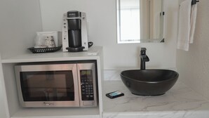 Un baño moderno con microondas integrado, cafetera y un lavamanos negro grande sobre encimera de mármol.