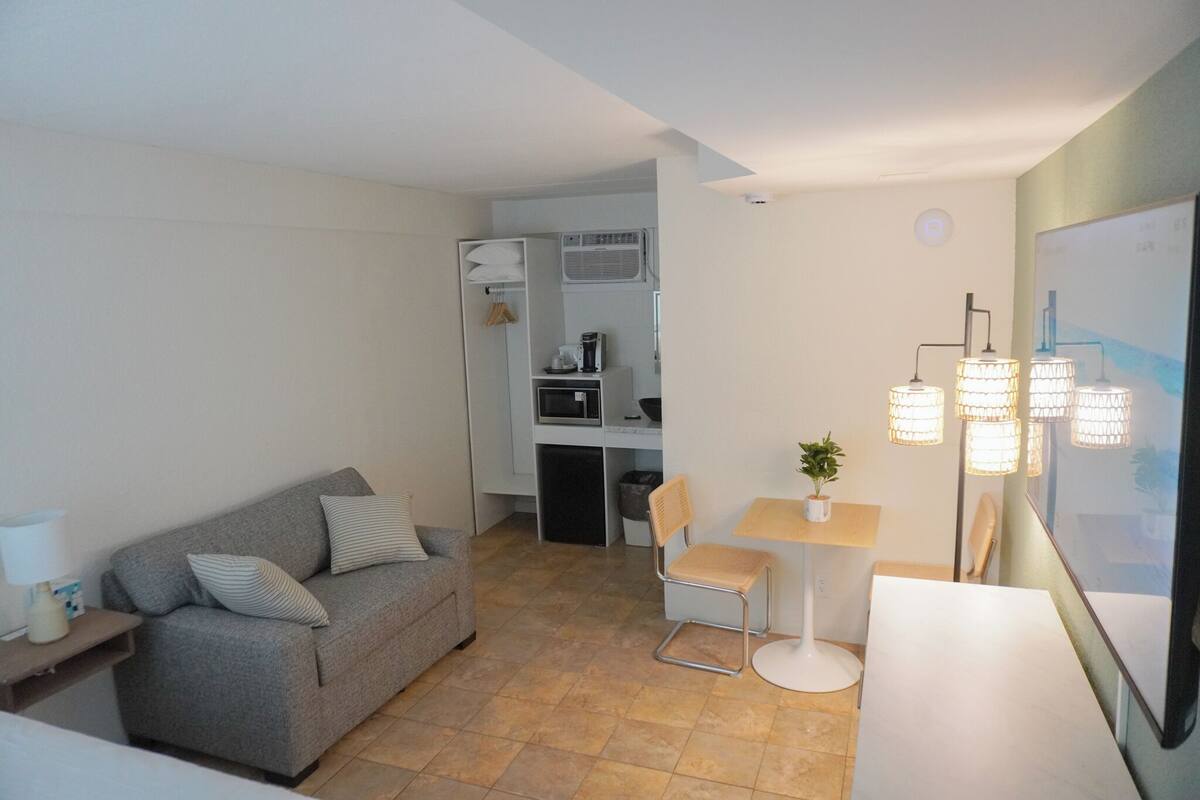 A compact living space with a grey sofa, a small dining table with two chairs, a white cabinet, and a wall-mounted air conditioner.