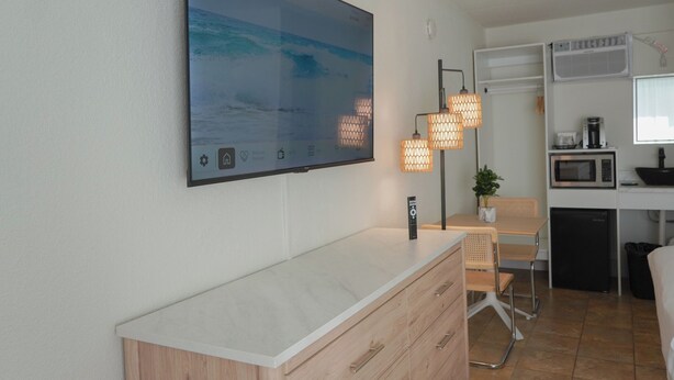 A modern room with a large flat-screen TV displaying a coastal scene, a compact kitchenette, a dining area with a table and chairs, and a small potted plant. - Pier Point (Wildwood)