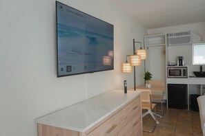 A modern room with a large flat-screen TV displaying a coastal scene, a compact kitchenette, a dining area with a table and chairs, and a small potted plant. - Pier Point (Wildwood)