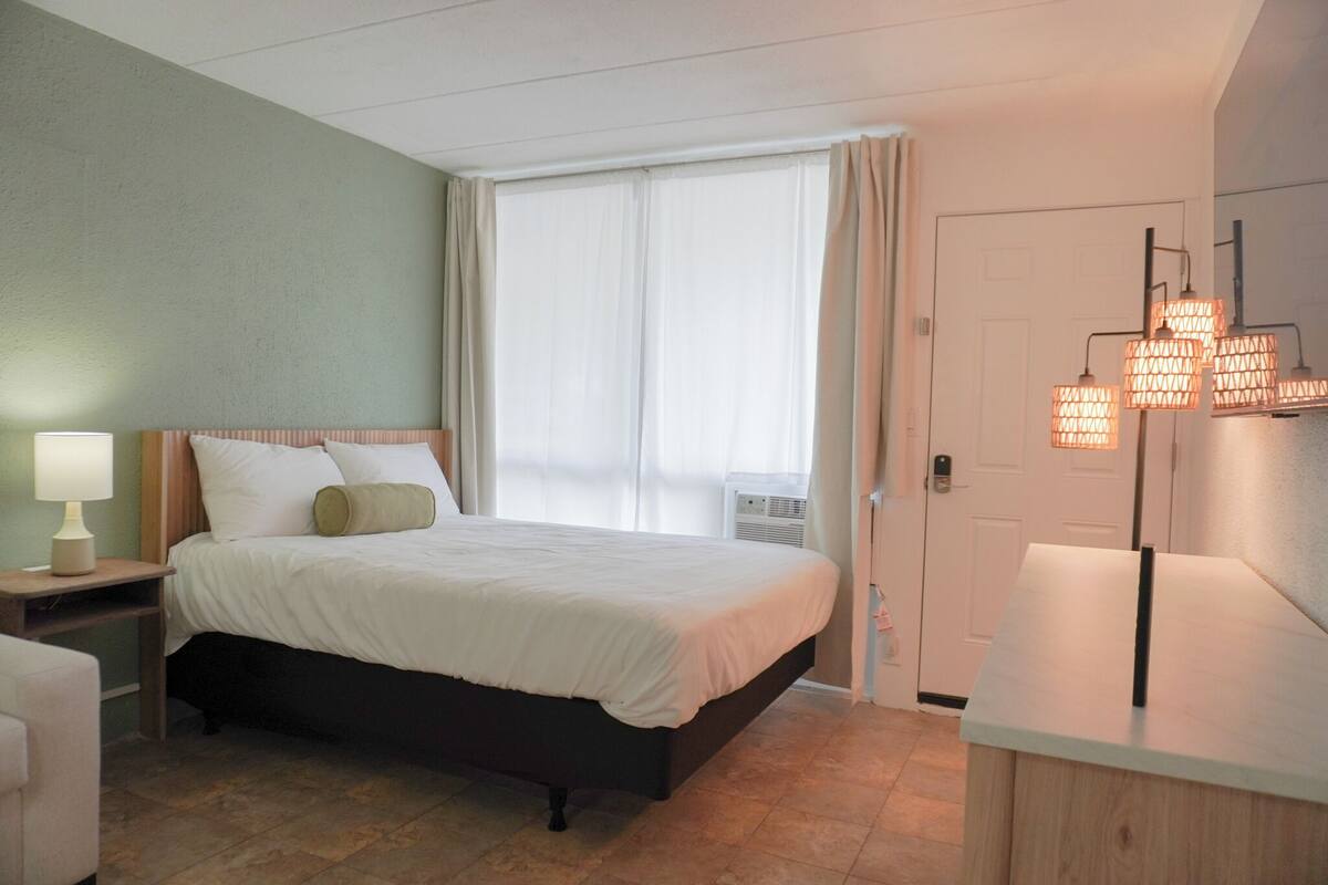 A hotel room with a bed, bedside table with a lamp, a wooden nightstand, a desk with a lamp, and a large window with curtains.
