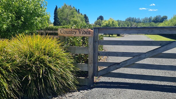 Ein hölzernes Tor mit einem Schild mit der Aufschrift 'Country Vibes', das zu einem Schotterweg führt.
