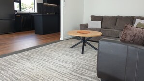 Una sala de estar moderna con un sofá gris, una mesa de centro redonda de madera y una cocina con armarios negros y un ventanal grande.