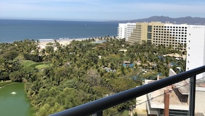 A view from a balcony overlooking a coastal area with a beach, palm trees, and a large hotel building. - 3-Bedroom Vidanta Grand Luxxe (Nuevo Nayarit)