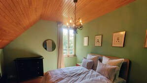 Une chambre avec un plafond en bois, un lit recouvert de linge de lit blanc, une commode, un miroir rond et des tableaux encadrés accrochés au mur.