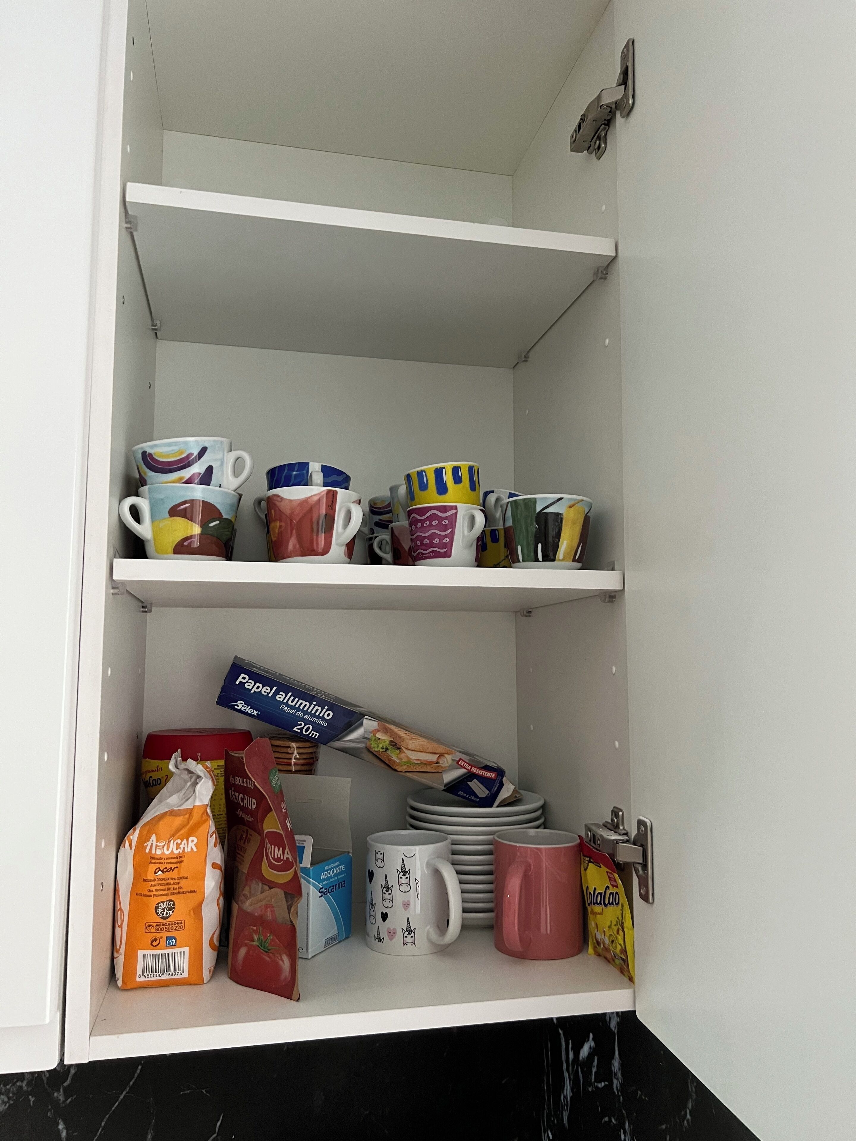 Un meuble avec des étagères contenant divers articles de cuisine, notamment des tasses colorées, des assiettes et des sacs d’épicerie.