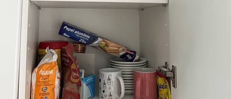 A cabinet with shelves containing various kitchen items, including colorful mugs, plates, and bags of groceries.