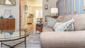 A living room with a glass coffee table, a beige sofa with patterned cushions, a wicker chair, and a view of the kitchen and dining area through an open door.