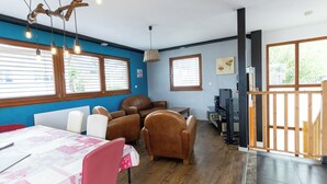 Un comedor con una mesa dispuesta para cuatro personas, rodeada de sillas de cuero. Hay dos ventanas con persianas, una lámpara en el techo y un televisor sobre un mueble.
