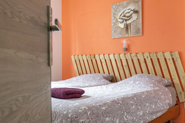 A bed with patterned bedding, a wooden headboard, a wall-mounted light fixture, and a framed artwork above the bed.