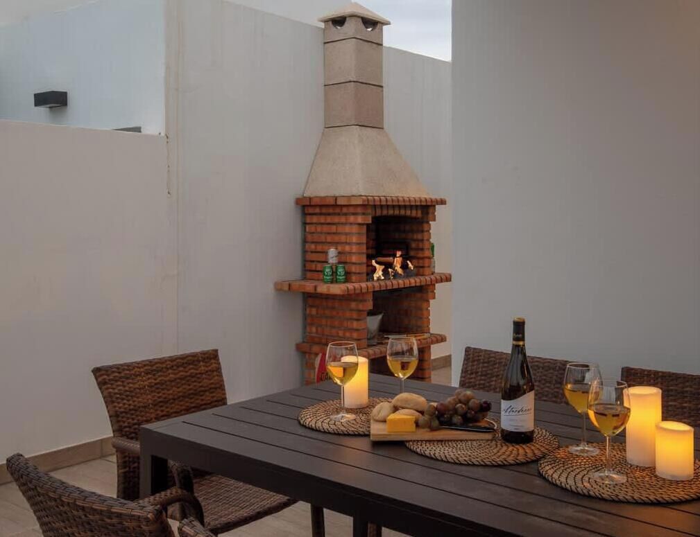 Un área de comedor al aire libre con horno de pizza de ladrillo, sillas de mimbre, una mesa con copas de vino, queso y una botella de vino.