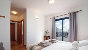A bedroom with a bed, a window with curtains, a bedside table with a plant, and a view of buildings outside.