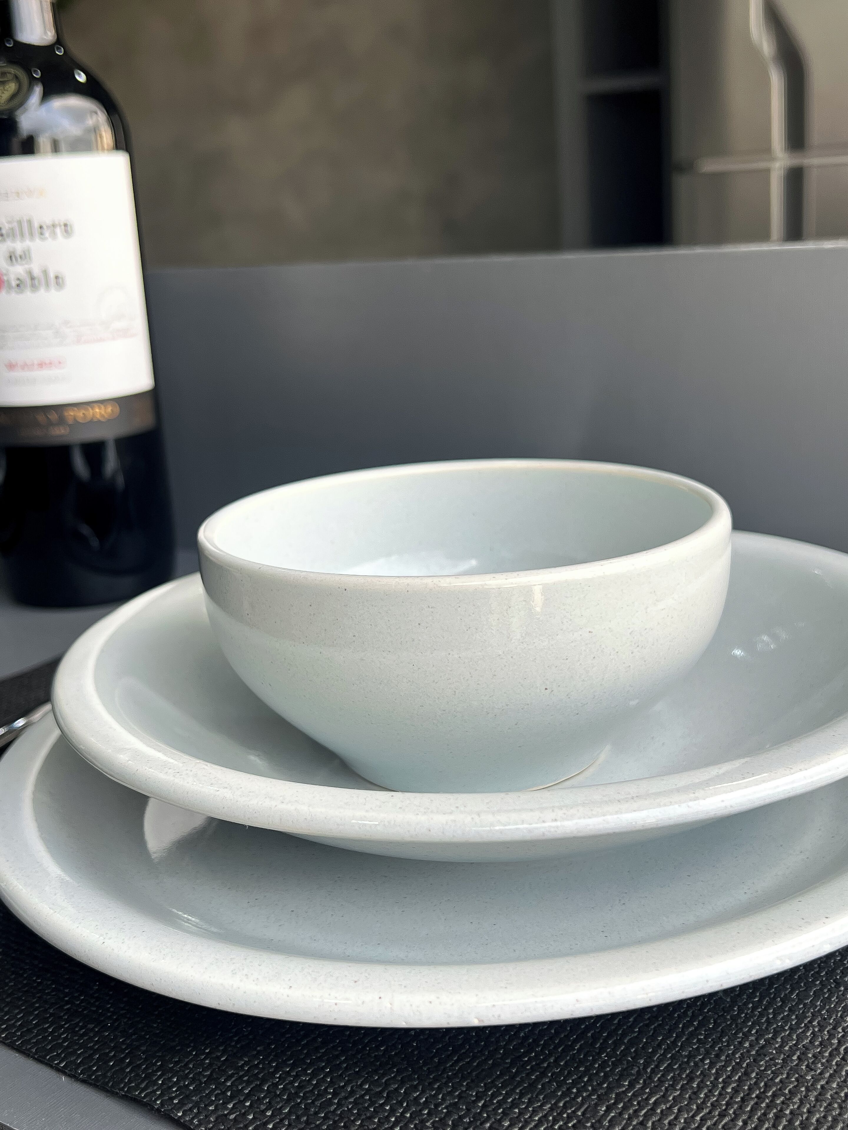 A white ceramic bowl on a matching plate, placed on a textured mat. A bottle of wine is visible in the background.
