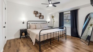 A bedroom with a bed, a ceiling fan, a bedside table, a window with curtains, and a mirror.